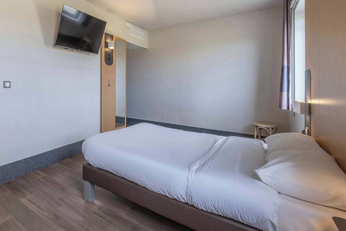 Habitación de hotel moderna con cama, televisor de pantalla plana en la pared, mesita de noche pequeña y ventana con cortinas.