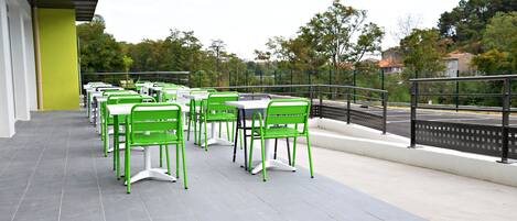 A terrace with green chairs and tables, a railing, and a view of trees and buildings.