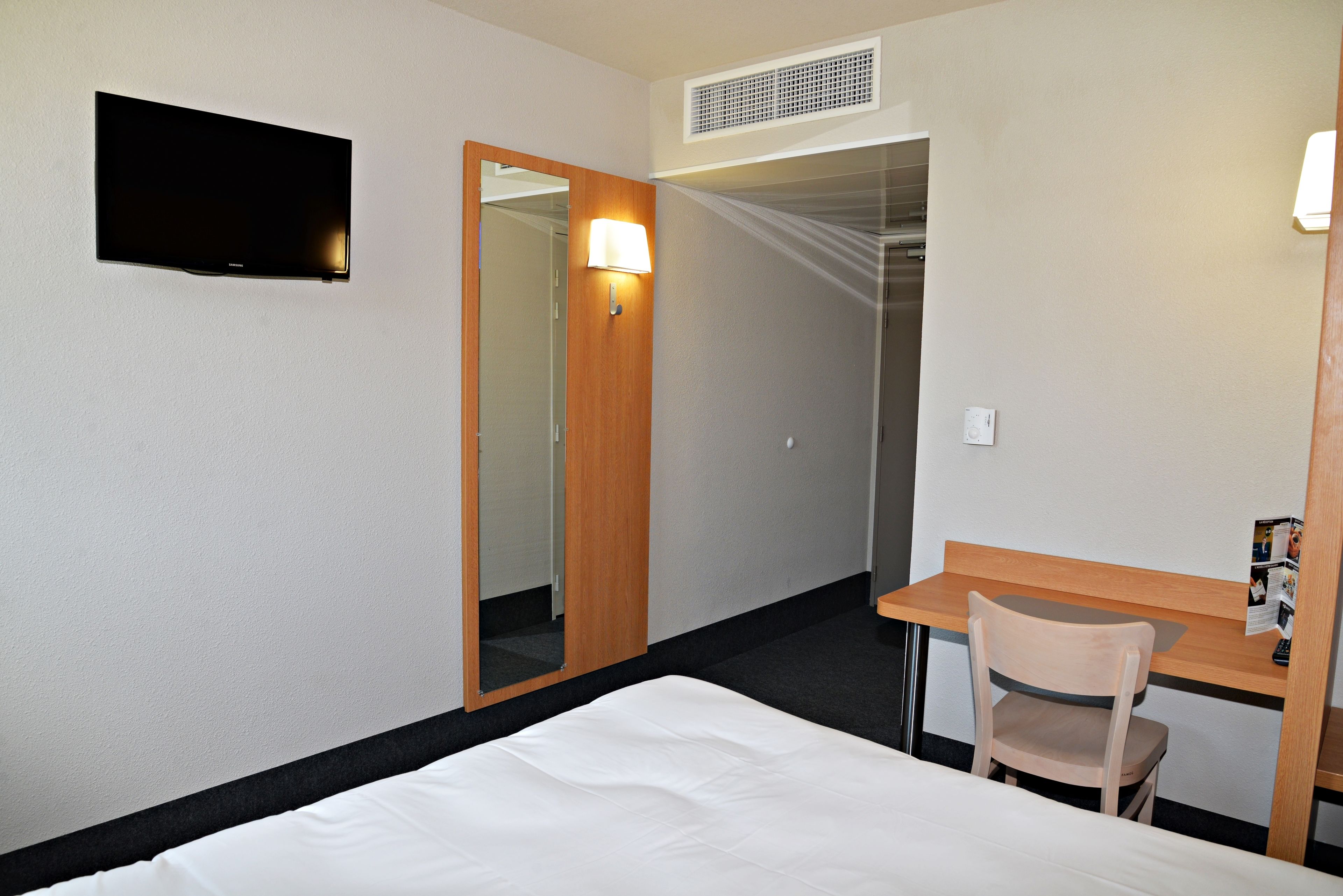Una habitación de hotel con cama, escritorio, silla, televisor y espejo.