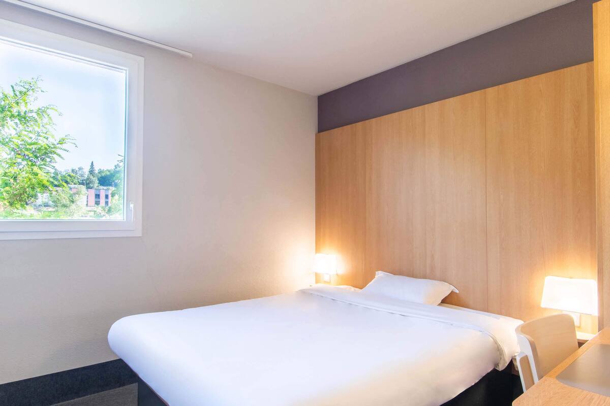 Ein Hotelzimmer mit einem großen Bett, zwei Nachttischlampen, einem hölzernen Kopfteil und einem Fenster mit Blick auf Grünflächen und Gebäude.