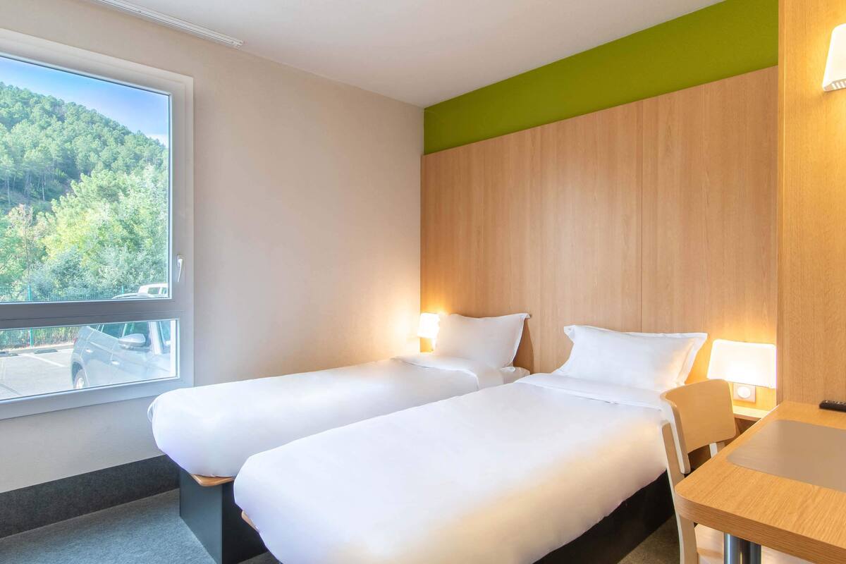 Ein Hotelzimmer mit zwei Einzelbetten, einem Holzkopfteil, einem Tisch und einem Fenster mit Blick auf eine bewaldete Landschaft.