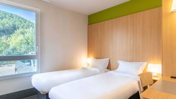 Ett hotellrum med två separata sängar, en sänggavel i trä, ett bord och ett fönster med utsikt över ett skogsområde.