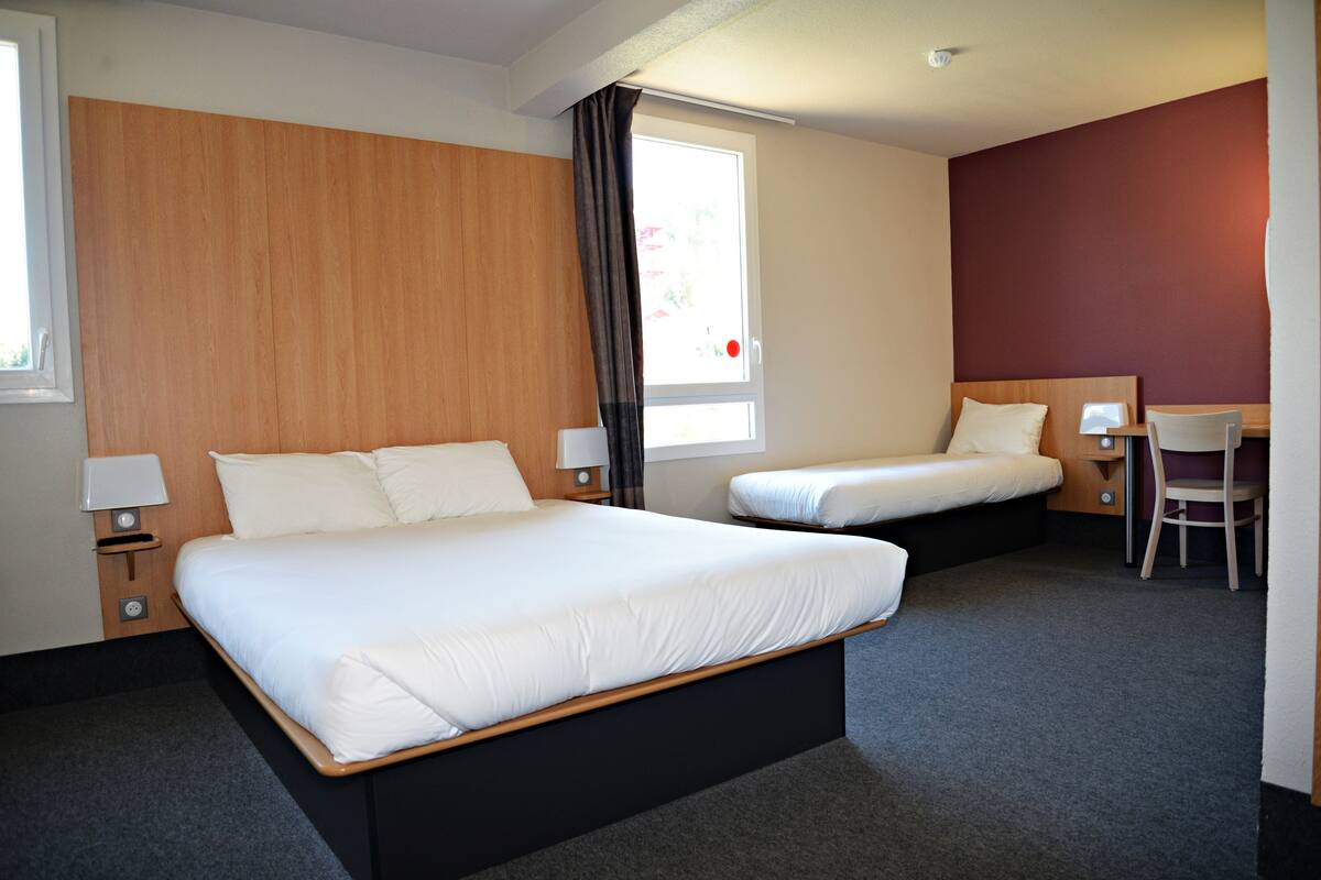 Ein Hotelzimmer mit zwei Betten, einem Schreibtisch, einem Stuhl, einem Fenster und einem Nachttisch mit Lampe.