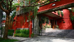 A red building with a covered entrance, a tree in front, and a stone pathway leading to the entrance. - Árbol de Fuego Eco-hotel, a green experience in the city (Antiguo Cuscatlán)