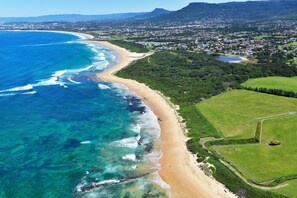 An aerial view of a coastal area with a beach, green fields, and a residential area. - Corrimal Beach Tourist Park (East Corrimal)