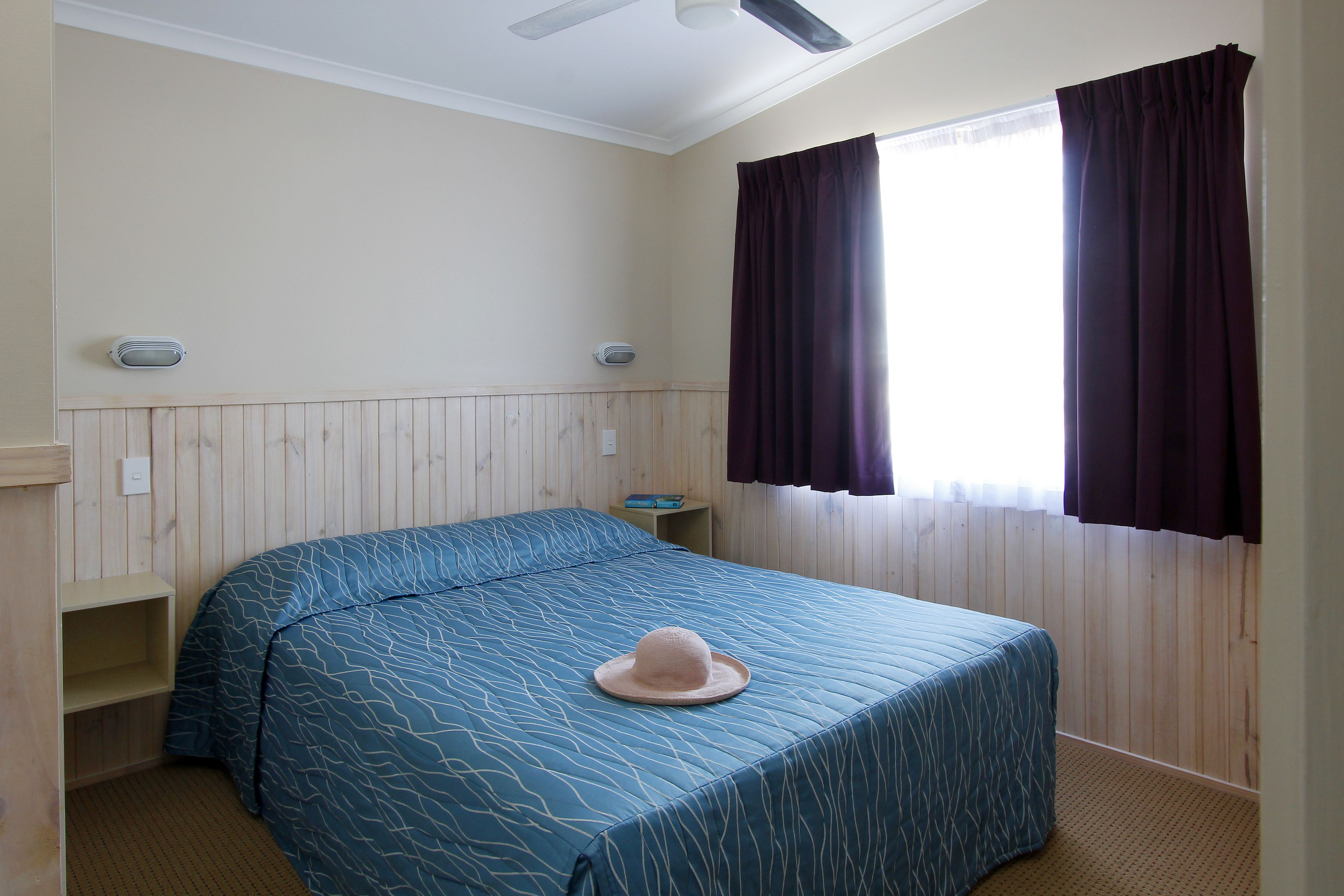 A bedroom with a bed, a hat on the bed, a window with curtains, a ceiling fan, and a shelf with a book.