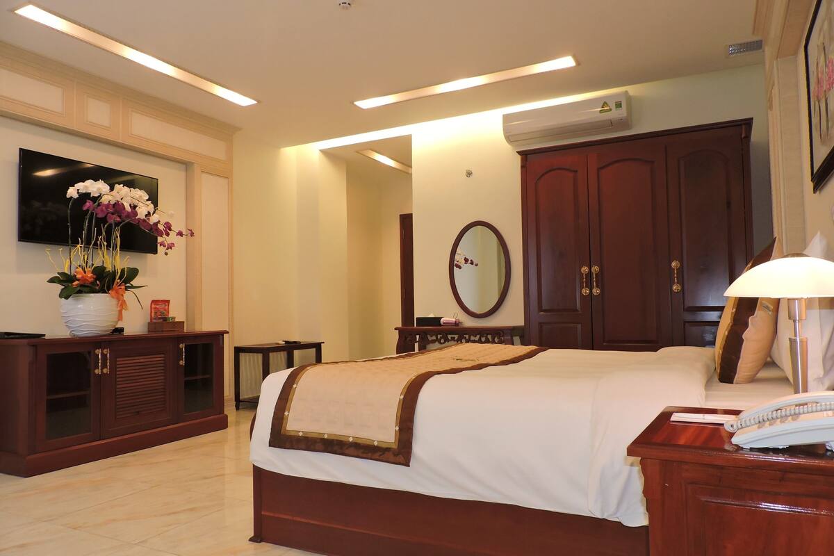 Habitación de hotel con una cama grande, un televisor montado en la pared, un armario de madera, un escritorio con lámpara y un espejo.