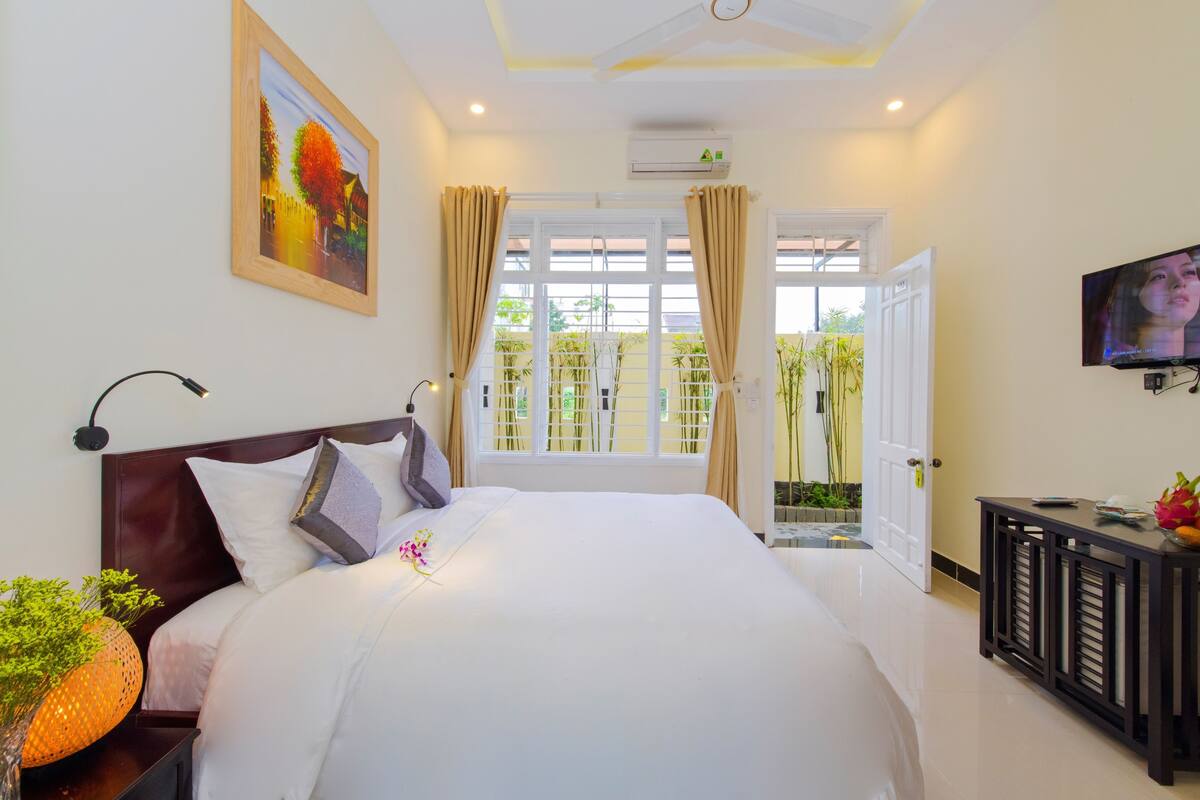 A hotel room with a large bed, a TV, a desk with a vase, and a window with curtains.