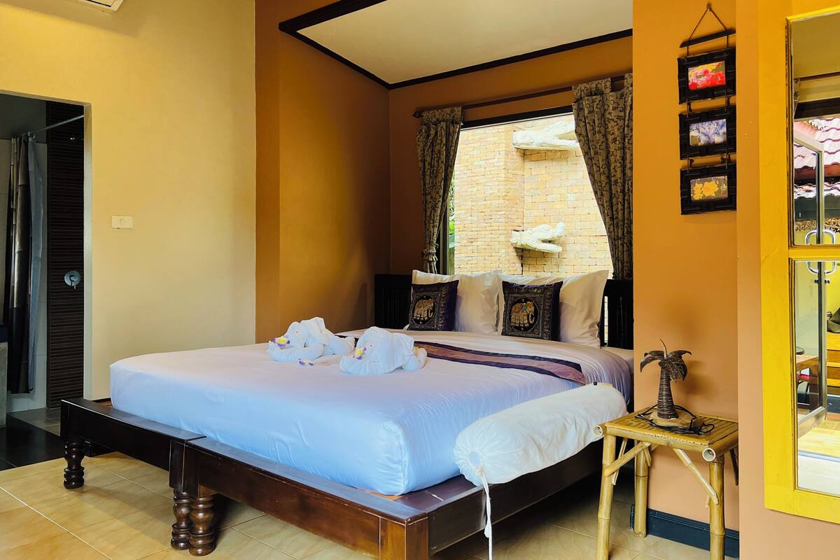 A bedroom with a bed, a window with curtains, a wall-mounted air conditioner, and a small table with a decorative item.