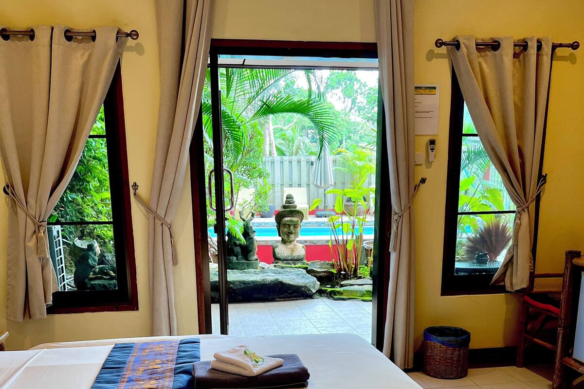 A hotel room with a bed, two towels, a view of a garden, and a statue outside.