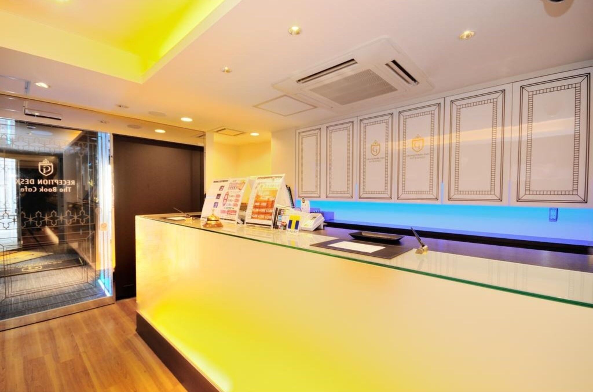 Un área de recepción con mostrador de cristal, conductos de aire acondicionado y decoraciones enmarcadas en la pared.