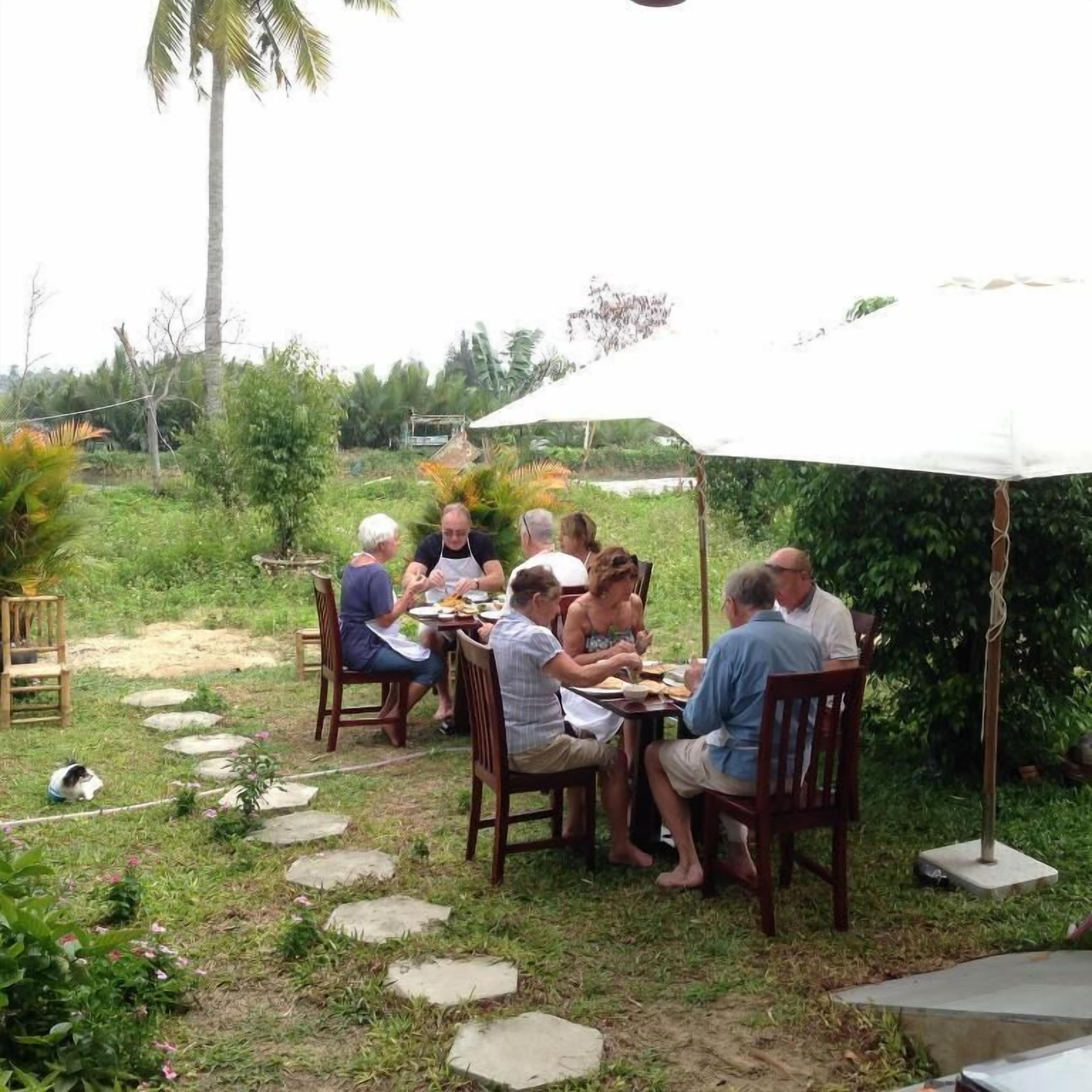 Un groupe de personnes est assis à une table à l’extérieur, sous un parasol blanc, et déjeune. L’ensemble est entouré de verdure et de chemins pavés.