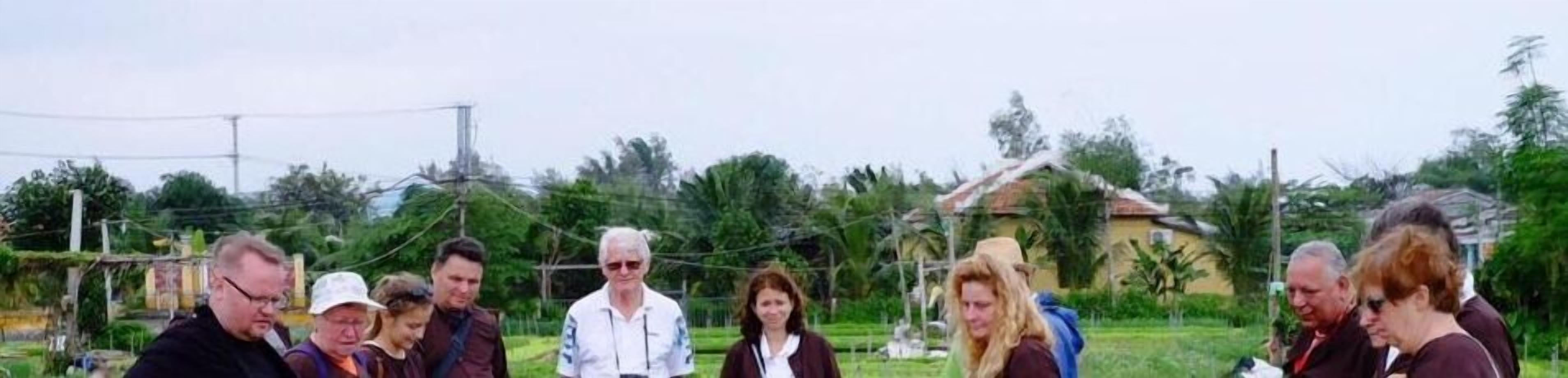 A group of people standing outdoors in a rural setting with greenery and a house in the background.