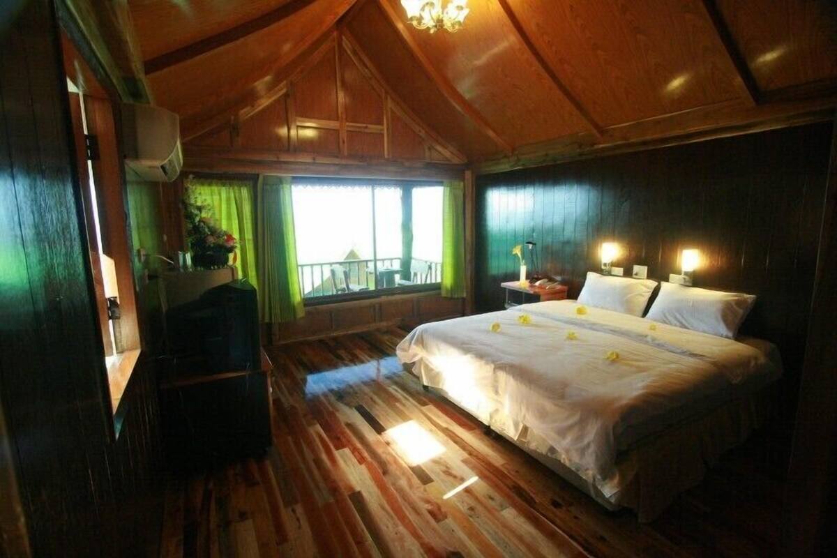 A wooden-floored bedroom with a bed, bedside lamps, and a view of the outdoors through a window.