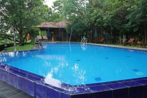 A rectangular swimming pool with a tiled edge, surrounded by trees and a covered seating area with chairs.