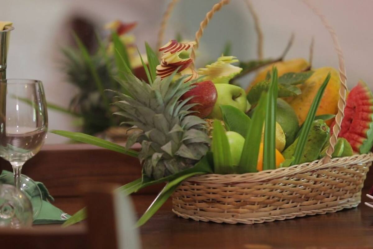 A dining table with a wicker fruit basket containing various fruits and a pineapple, a wine glass, and a red napkin.