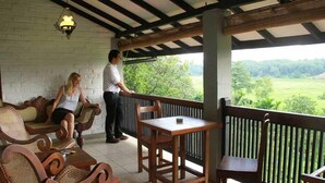 A balcony with wicker furniture and a table, overlooking a lush green landscape. - Lake Serenity Boutique Hotel (Kuruwita)