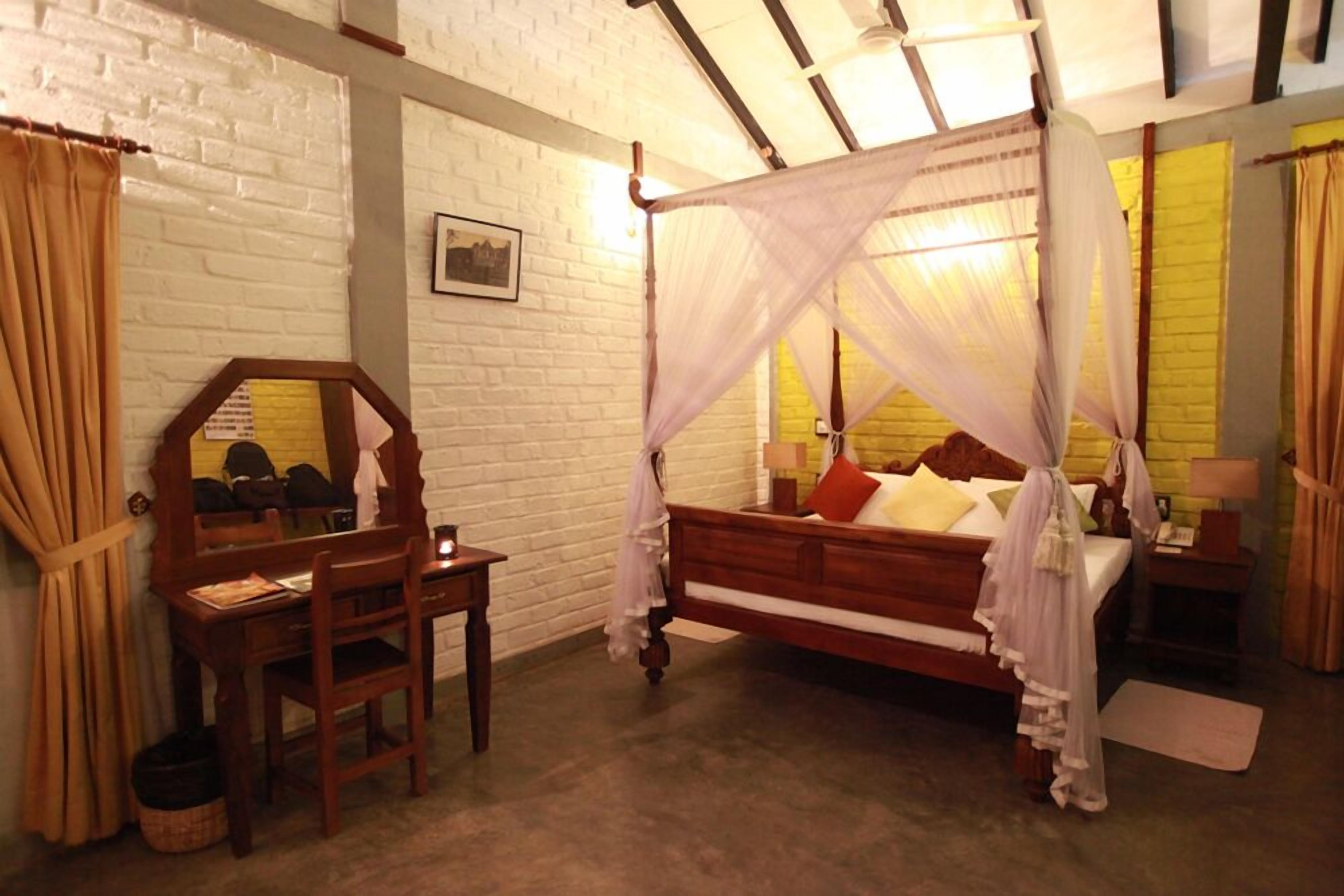 A bedroom with a four-poster canopy bed, a wooden desk with a chair, a mirror, and a bedside table with a lamp.