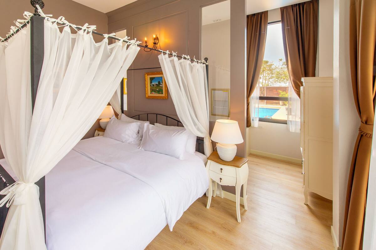 A bedroom with a four-poster bed, a bedside table with a lamp, and a view of an outdoor swimming pool.