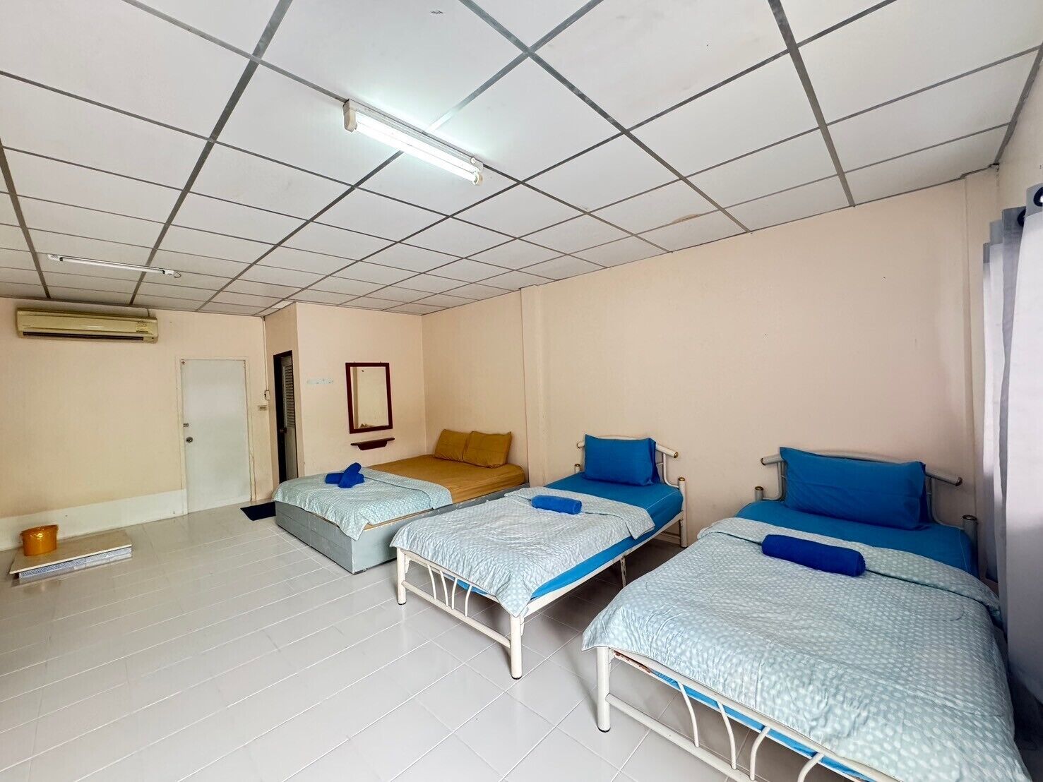 A room with three single beds, a white air conditioning unit, a door, and a mirror on the wall.