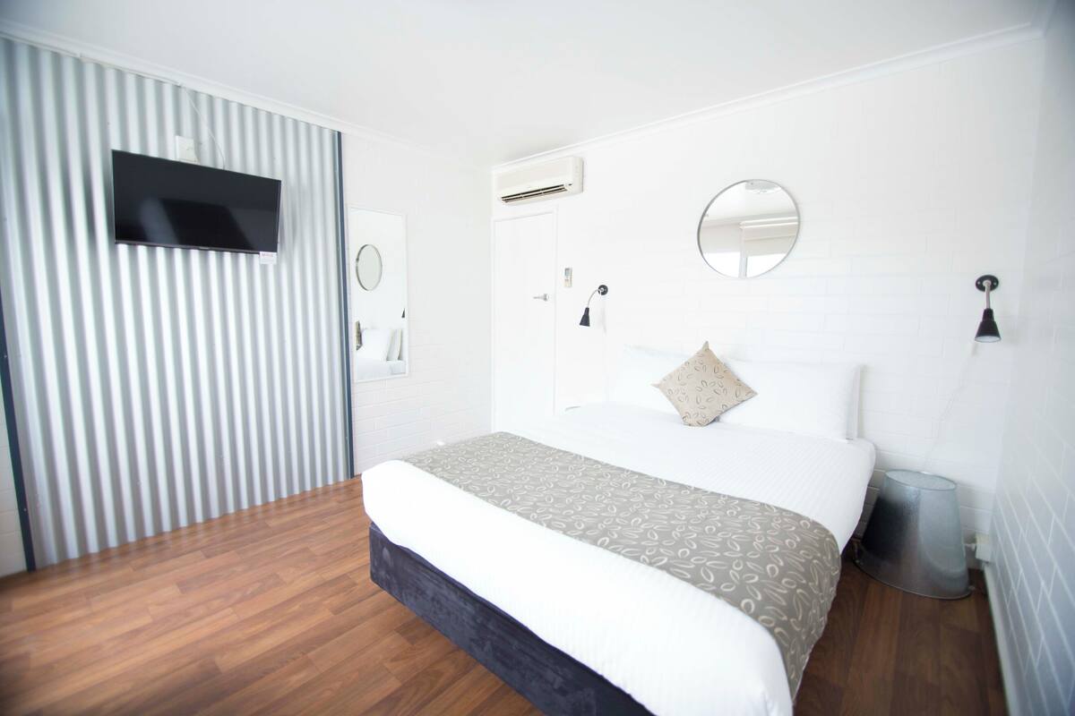 A modern bedroom with a large bed, a TV mounted on the wall, an air conditioning unit, and a mirror.