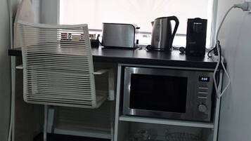 Cocina compacta con microondas, tostadora, hervidor y cafetera sobre una encimera. Un pequeño estante abajo contiene tazas y un hervidor.