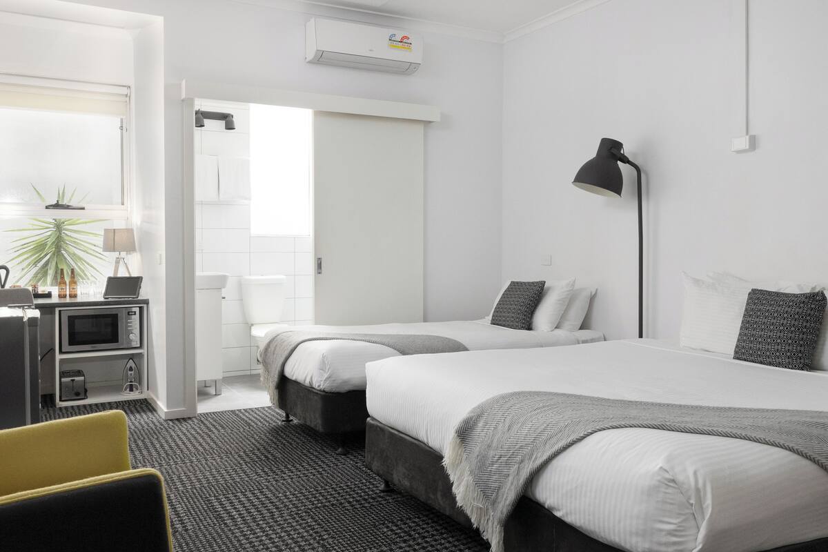 A modern hotel room with two beds, a desk with a laptop, a microwave, and a small kitchenette.