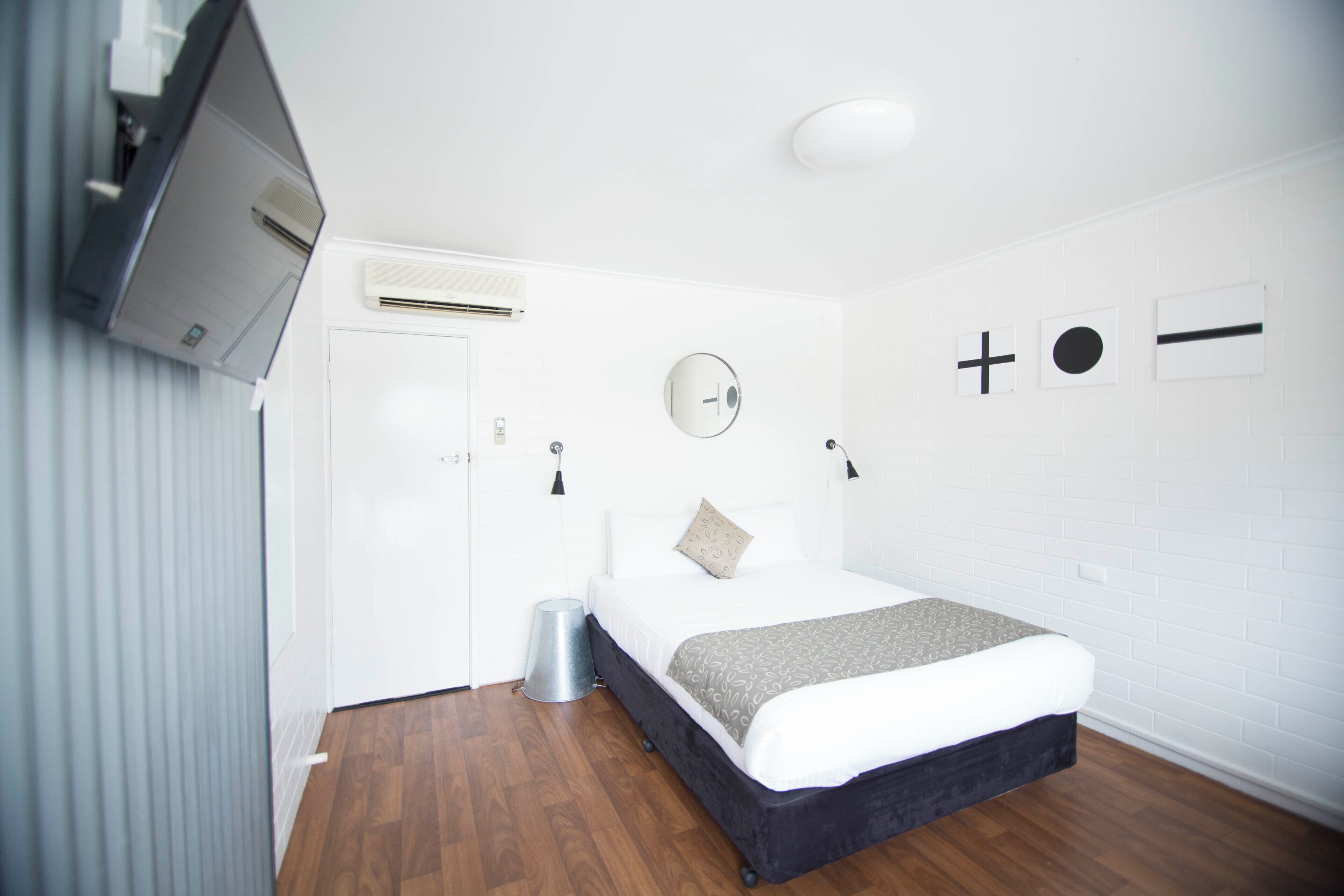 A modern bedroom with a bed, a TV mounted on the wall, a mirror, and a small trash bin.