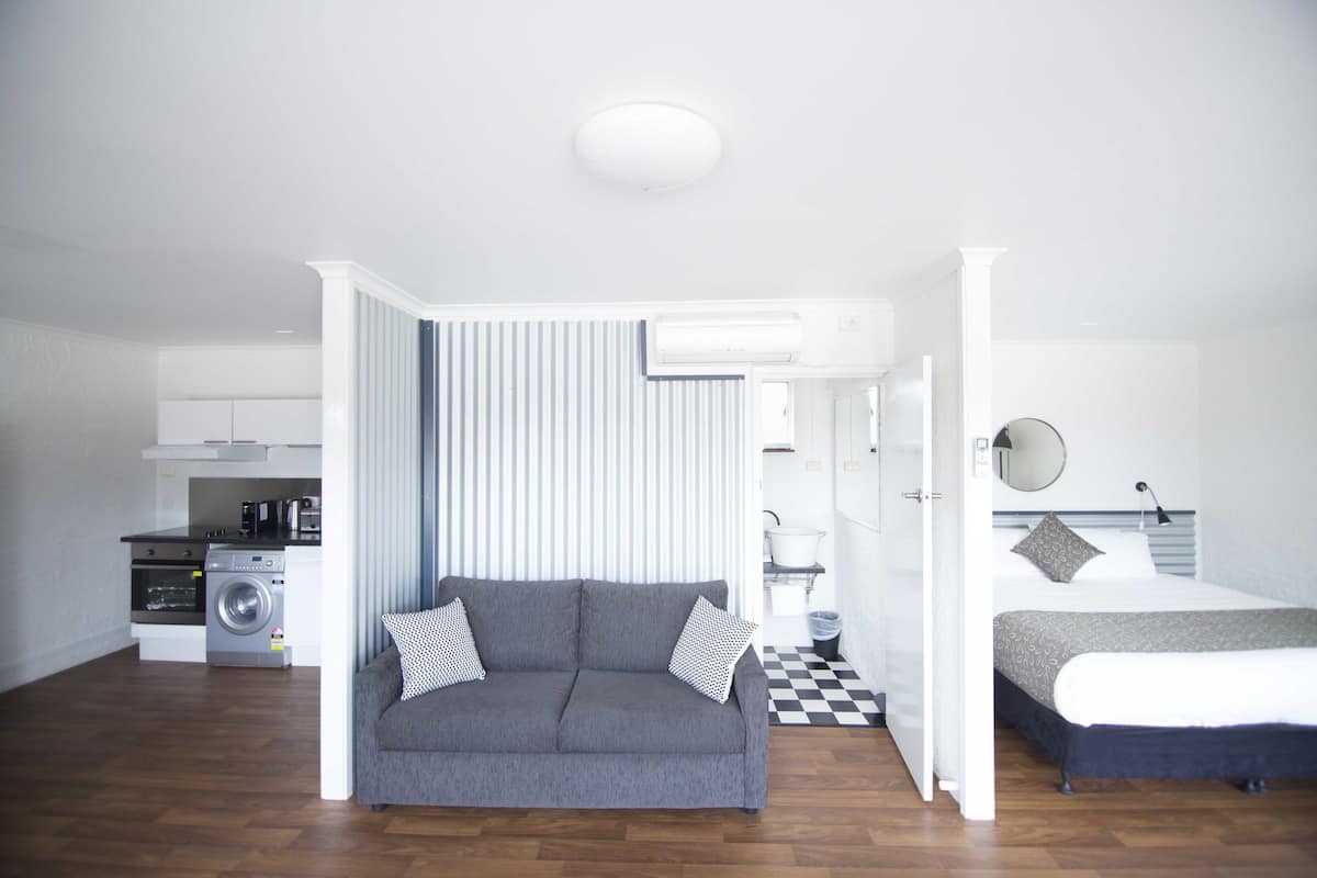 A compact hotel room with a grey sofa, a bed with patterned bedding, a small kitchen area with a washing machine, and a checkered floor.