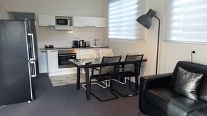 A compact kitchen with a refrigerator, microwave, and toaster, a dining table with chairs, a black leather sofa, and a floor lamp. - Siesta Central Apartments (Sale)