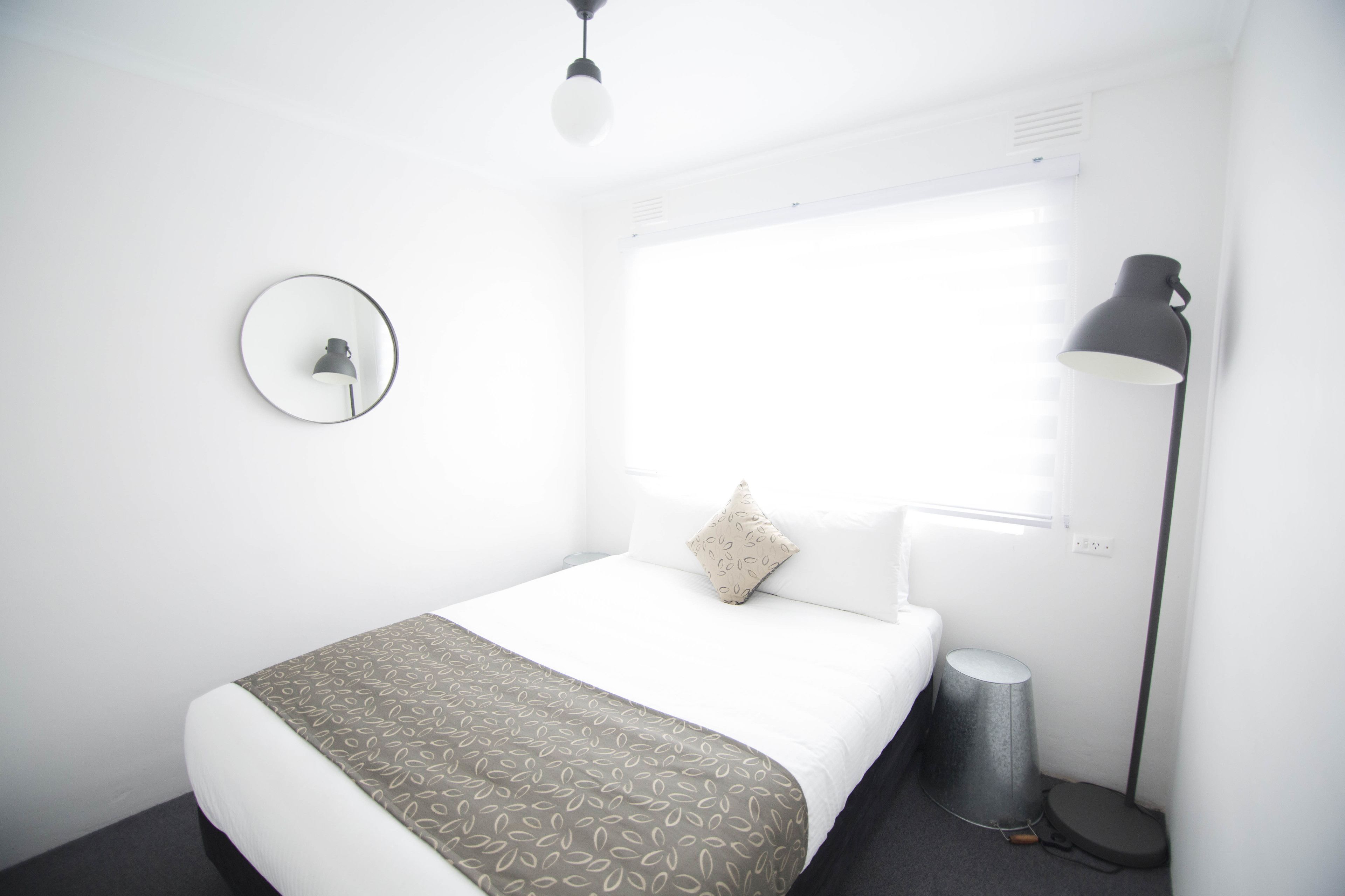 A hotel room with a bed, a round mirror, a bedside lamp, and a window with blinds.