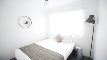 A hotel room with a bed, a round mirror, a bedside lamp, and a window with blinds.