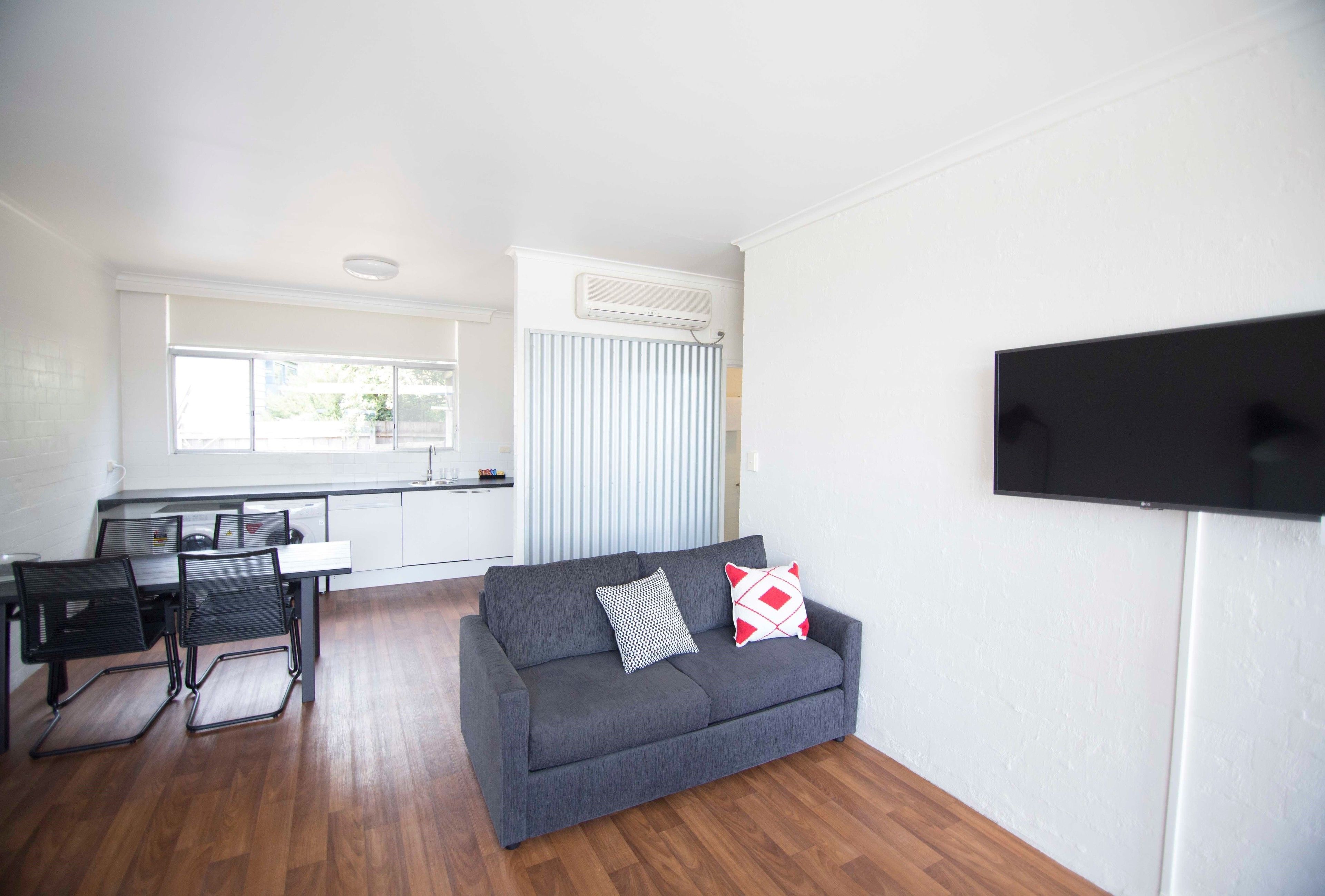 A modern living room with a grey sofa, two black chairs, a flat-screen TV, and a kitchen area with a window and blinds.