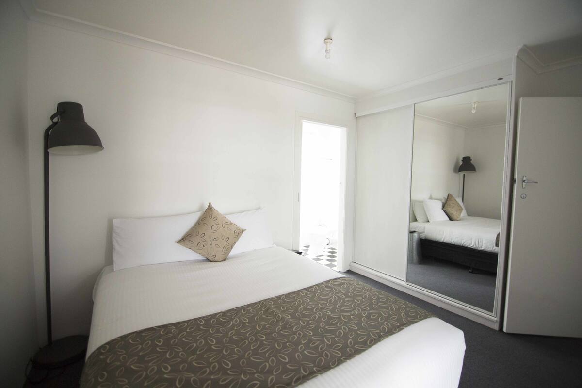 A hotel room with a bed, a bedside lamp, a mirror, and a door.