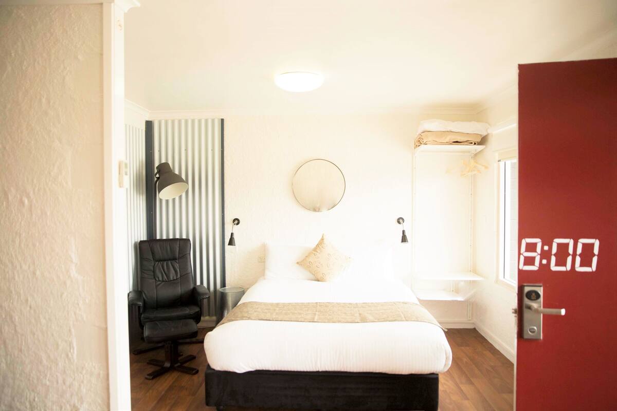 A hotel room with a bed, a chair, a mirror, a clock, and a door.