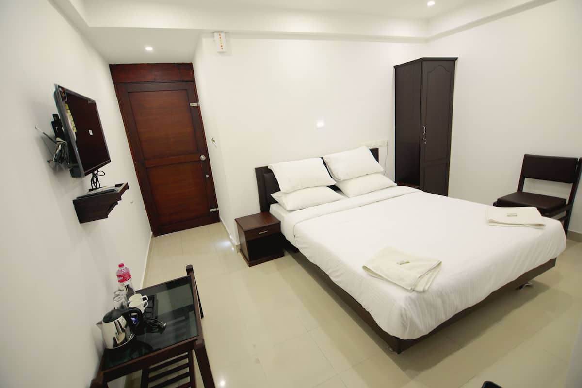 A hotel room with a large bed, a bedside table, a chair, a wardrobe, a TV, and a small table with a kettle and a bottle.