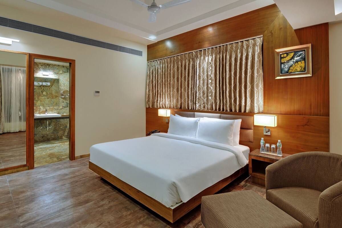 A hotel room with a large bed, a wooden headboard, a nightstand with water bottles, a chair, and a mirror.