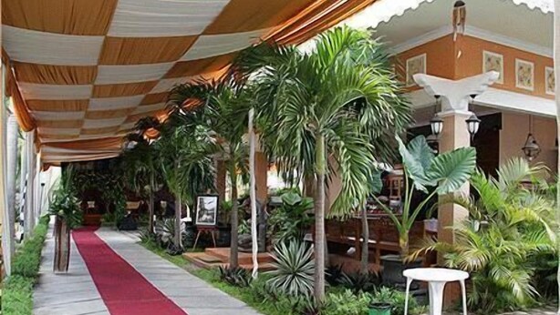 A covered walkway with a red carpet, surrounded by tropical plants and a white table. - Patria Garden Hotel (Blitar)