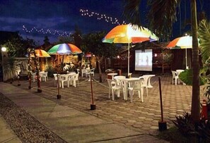 Un espace extérieur aménagé en terrasse, avec des chaises et des tables en plastique blanc, des parasols colorés et un écran de projection.