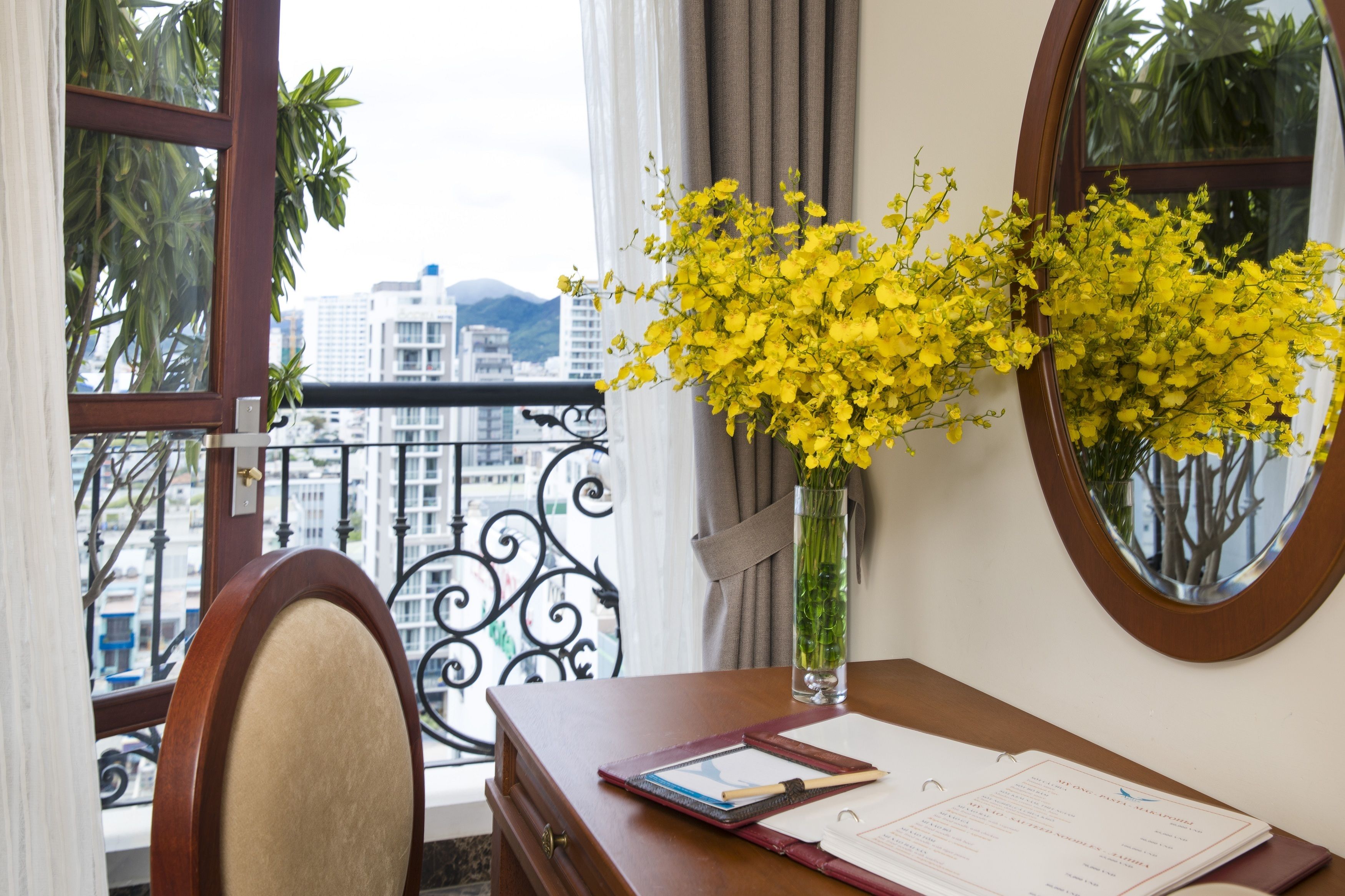Une pièce avec un bureau, un vase rempli de fleurs jaunes, une fenêtre donnant sur des immeubles et un miroir accroché au mur.