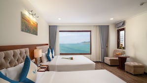 Habitación de hotel con un ventanal grande con vista al mar, una cama con ropa de cama azul y blanca, una mesita de noche con una lámpara y una pequeña mesa con un florero.