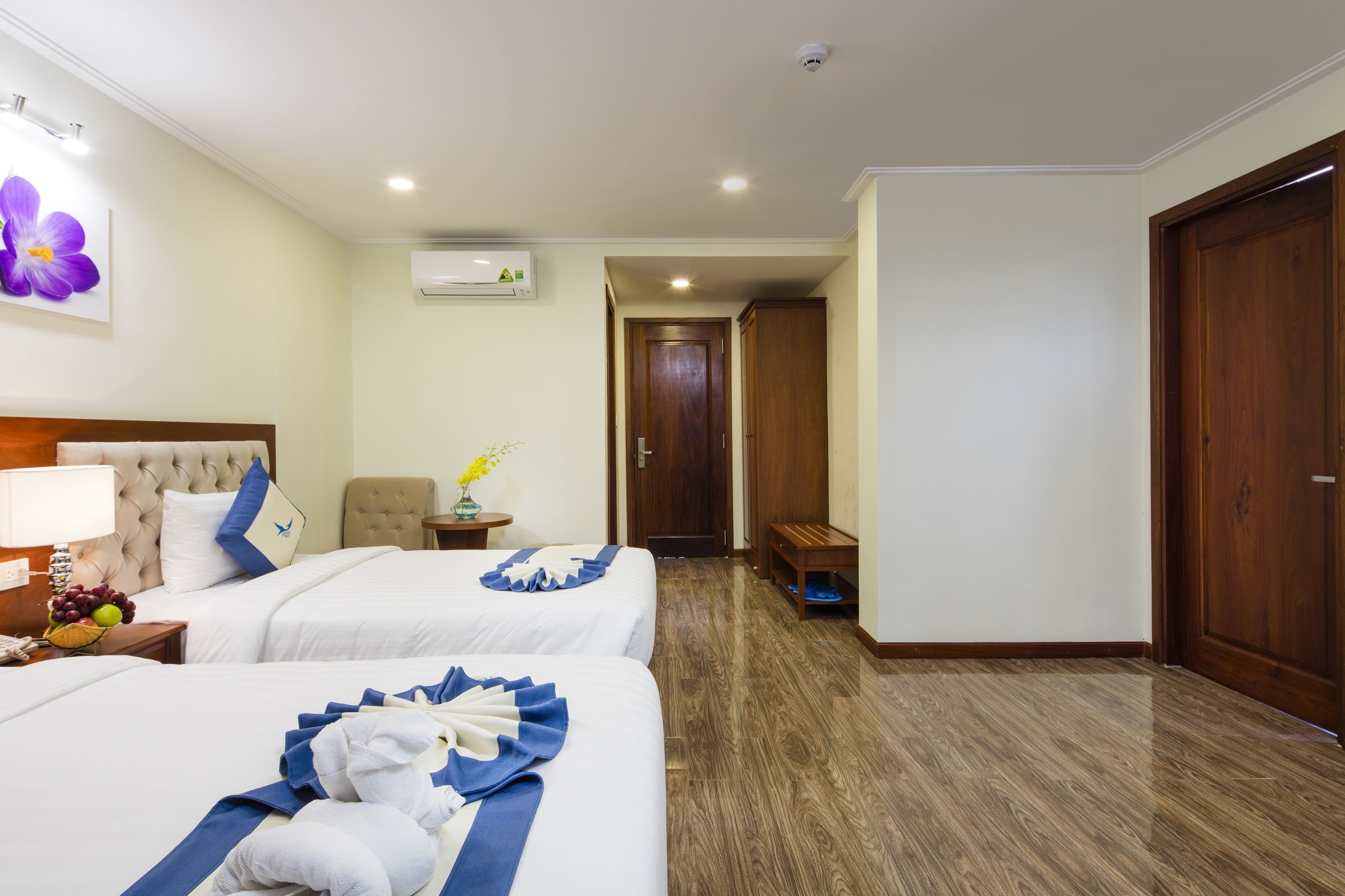 A hotel room with two beds, a wooden floor, a bench, a small table with a vase, and a wall-mounted air conditioner.