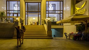 Der Eingang des Apus Hotels mit einer zur Eingangstür führenden Treppe, großen Fenstern und einem Schild über den Türen.