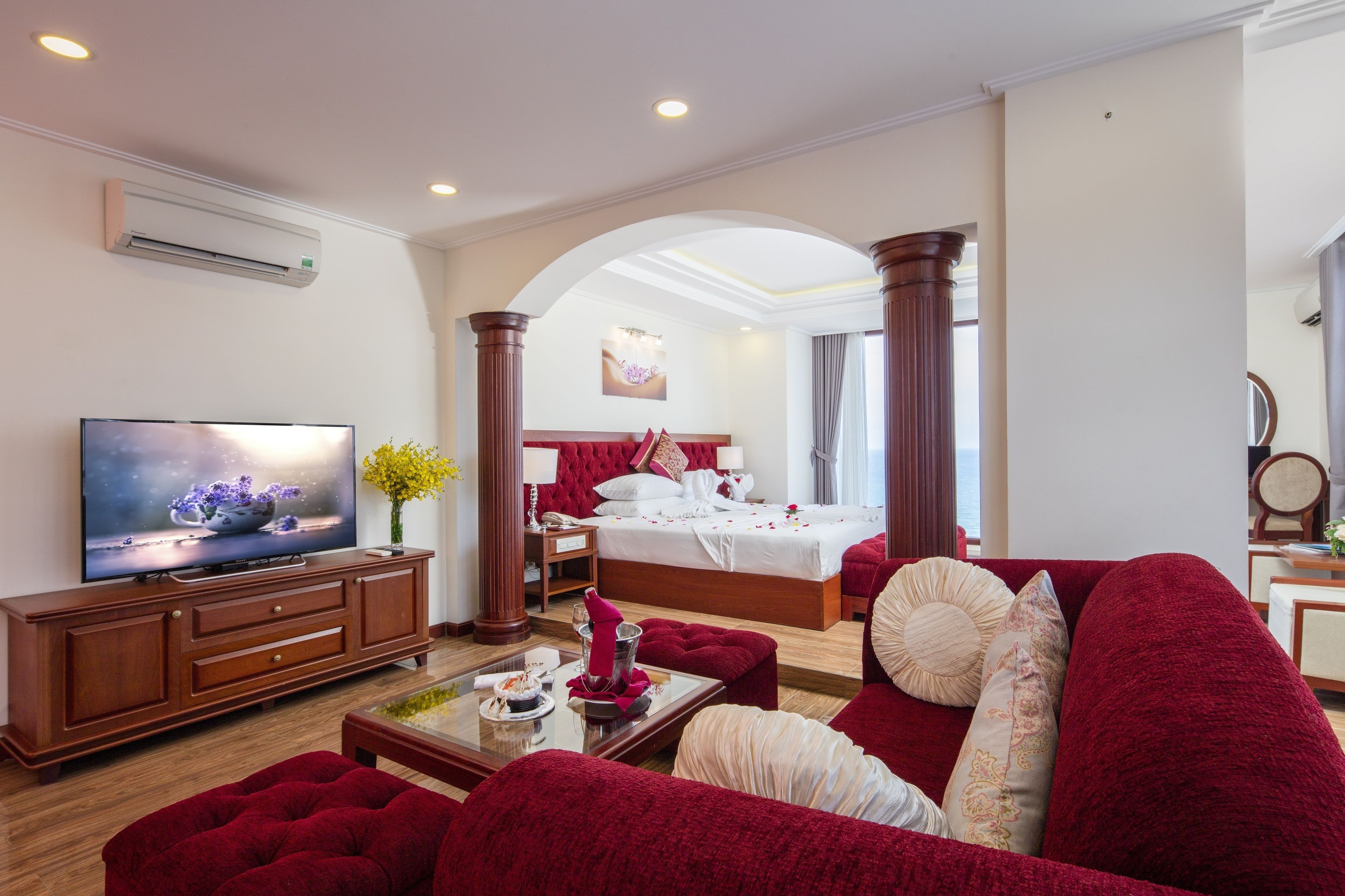 Habitación de hotel con una televisión de pantalla plana grande, cama con ropa de cama roja, sofá rojo y una mesa de centro de madera.