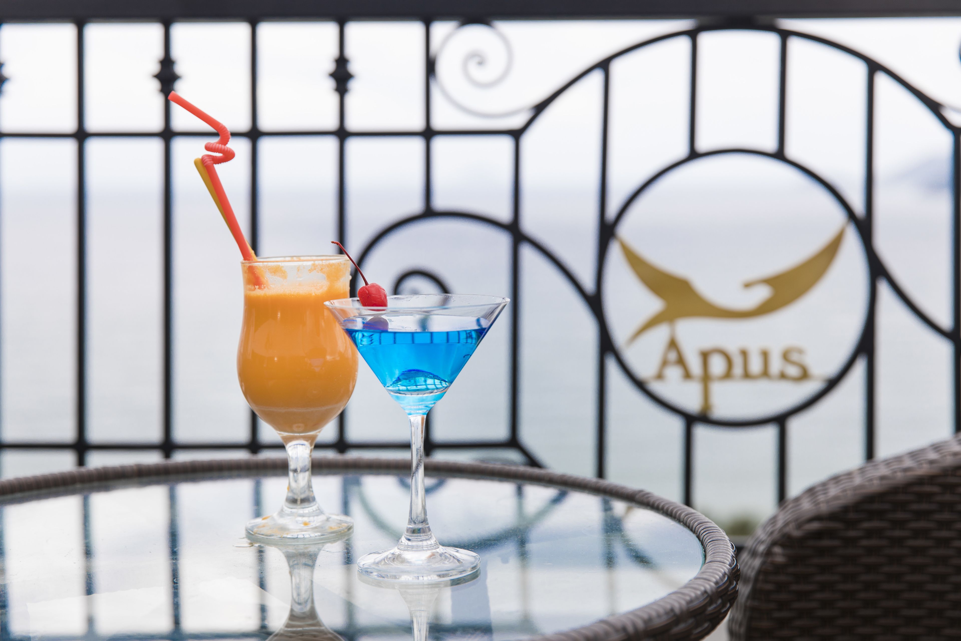 Two cocktails on a glass table with a wrought iron gate and a sign in the background.