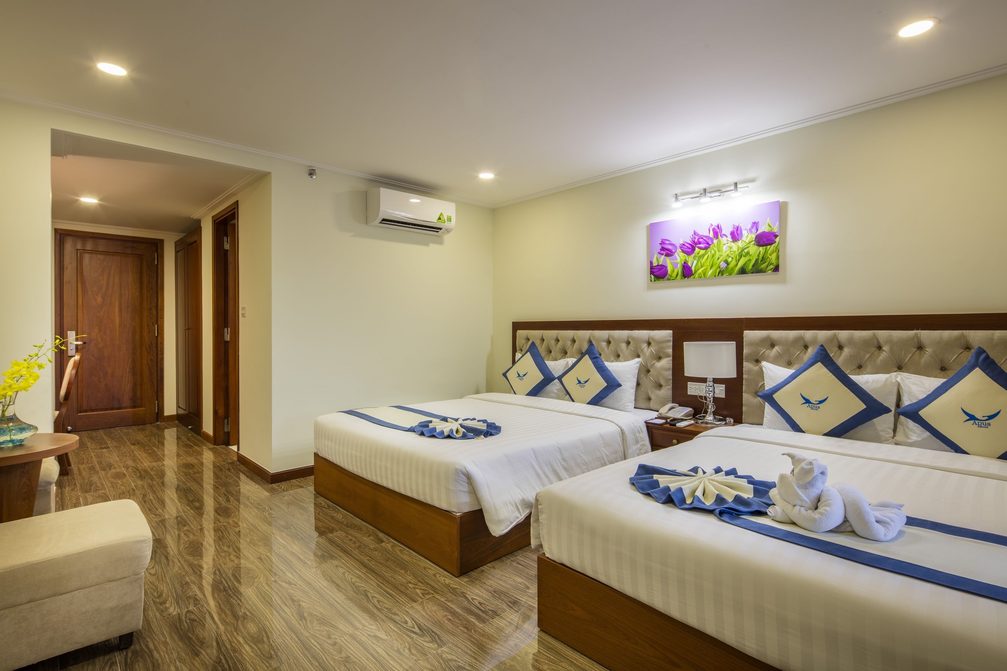A hotel room with two beds, a wooden floor, an air conditioning unit, a painting of flowers, and a desk with a vase of flowers.