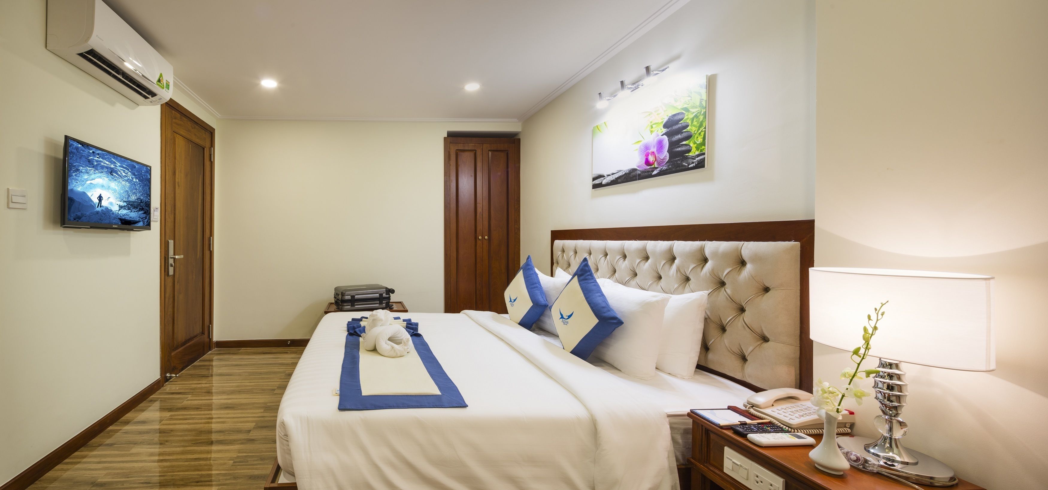 A hotel room with a large bed, a television, a painting, and a desk with a lamp.