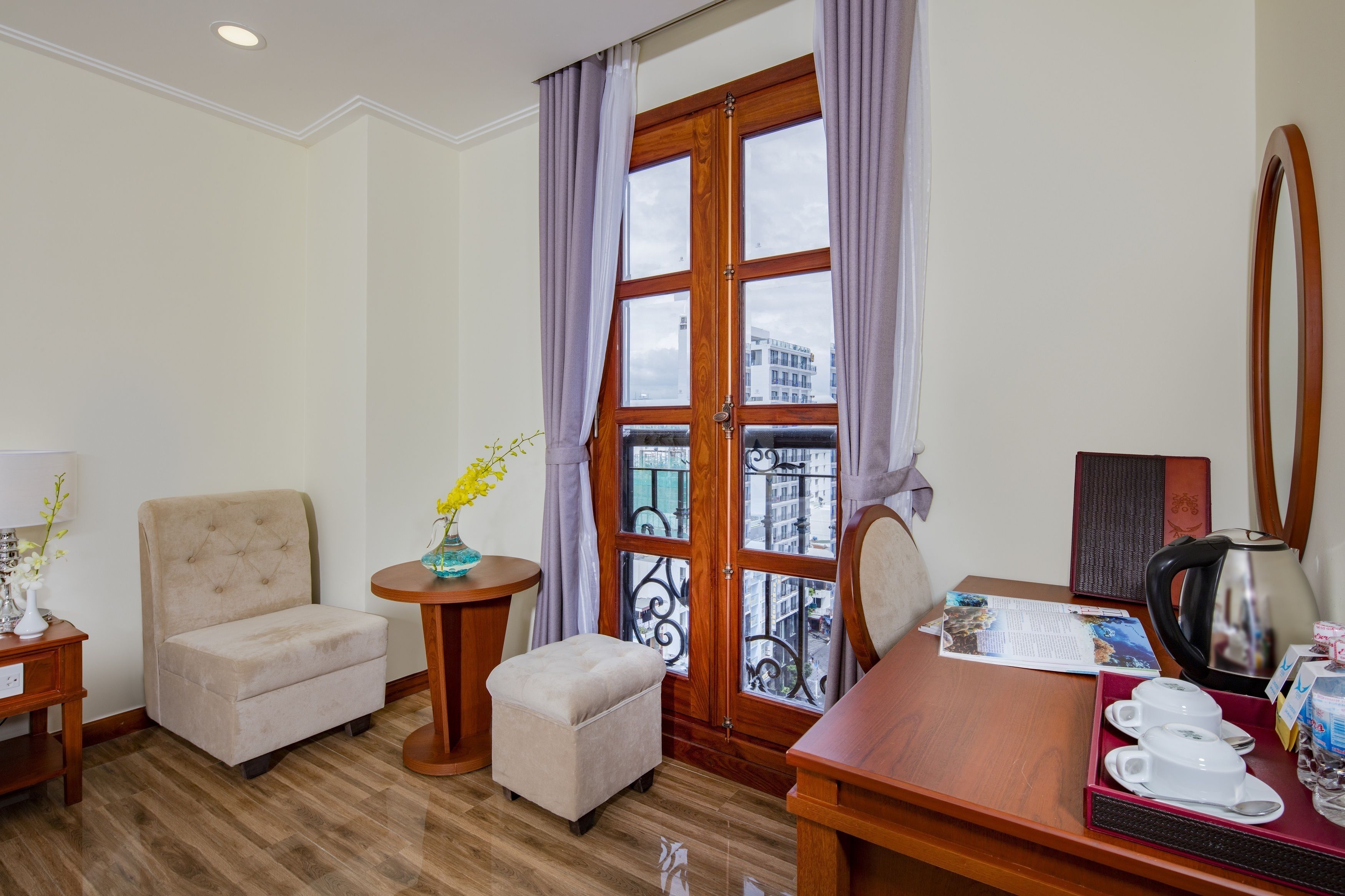 A hotel room with a wooden desk, a chair, a small ottoman, a side table with a vase, and a window with curtains.