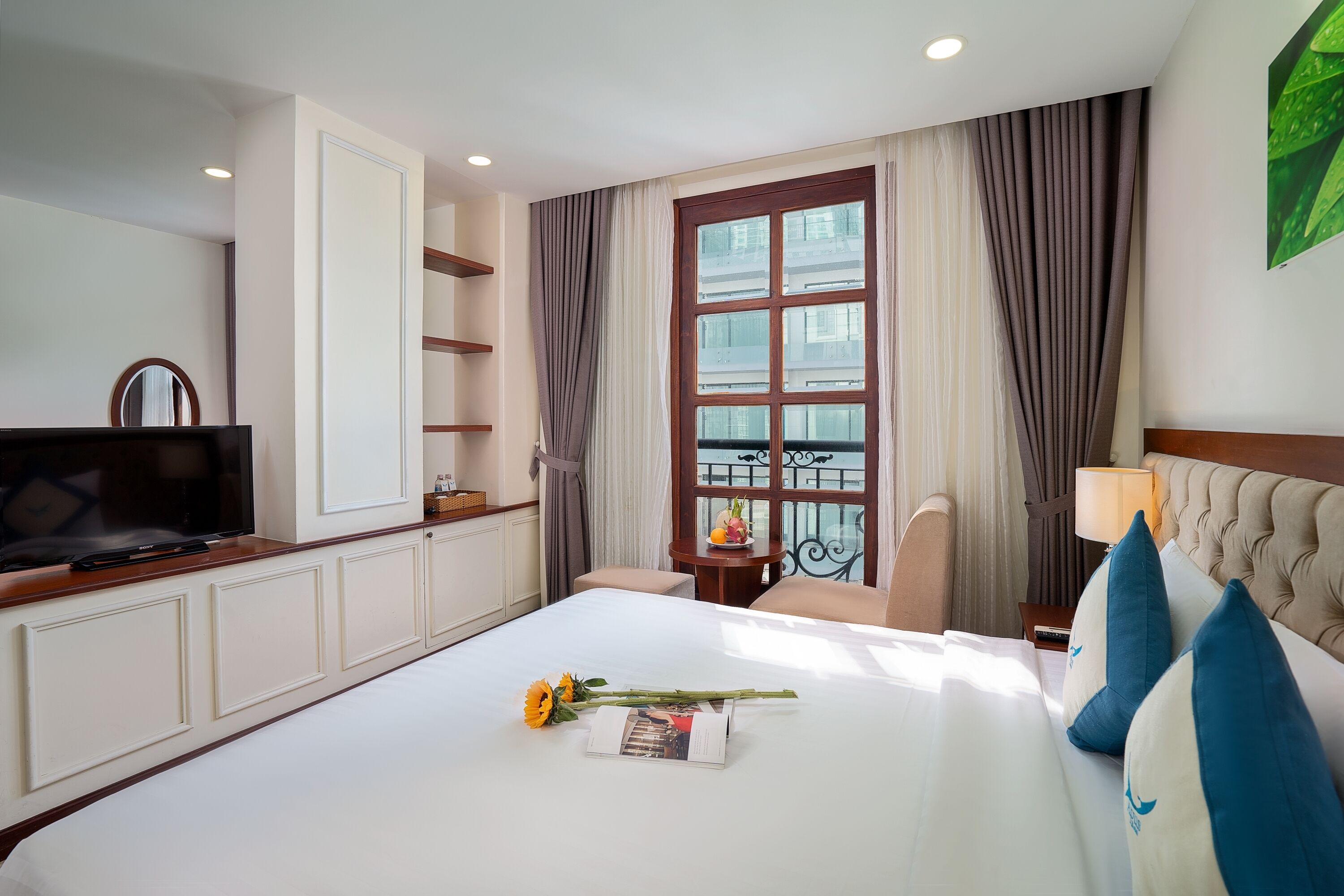 Une chambre d’hôtel avec un grand lit, une télévision, une fenêtre avec des rideaux, une lampe et une petite table sur laquelle se trouve un vase.