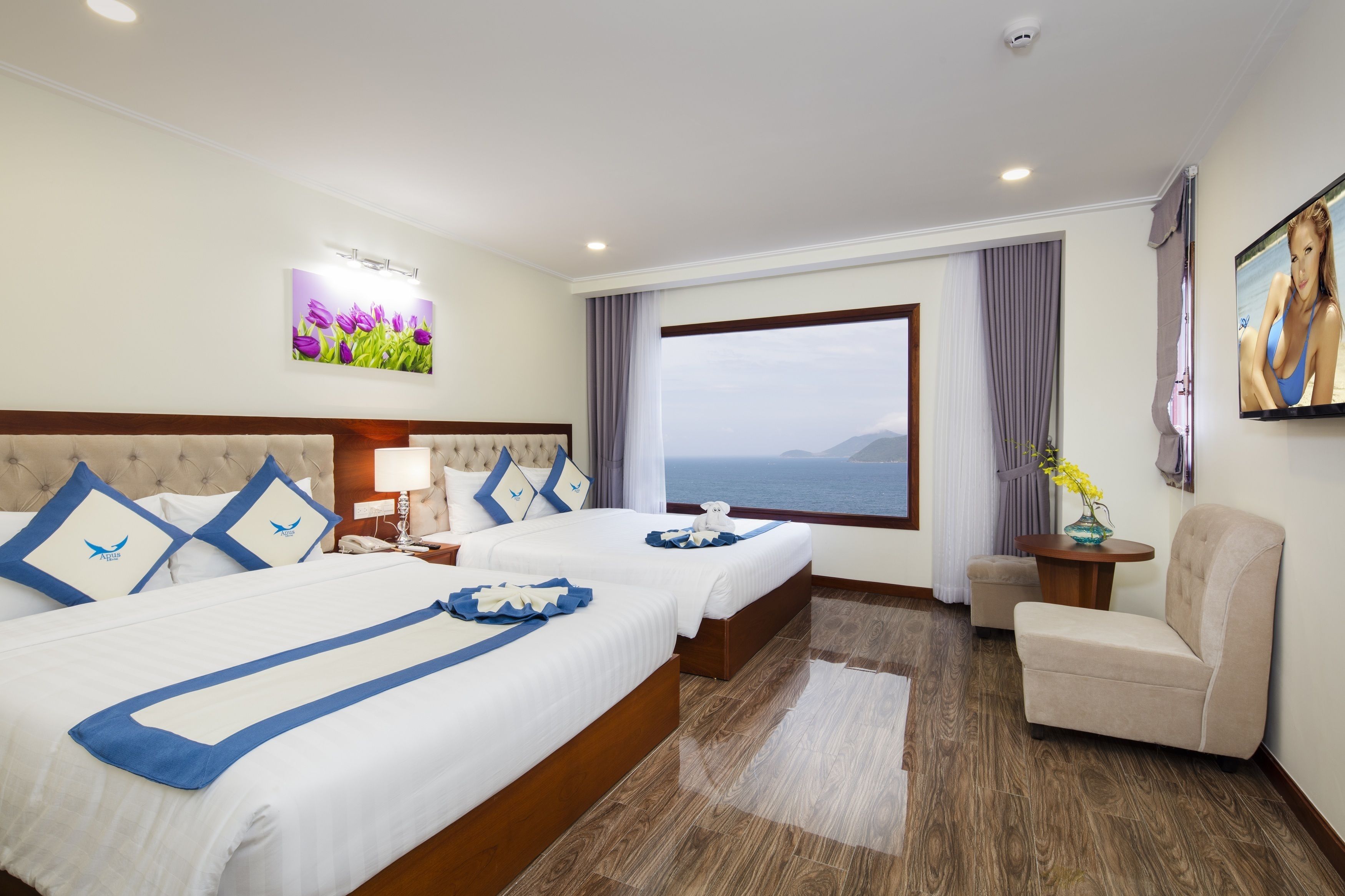 Habitación de hotel con dos camas, vista al mar, televisión y una pequeña mesa con un florero.